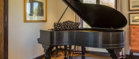 A grand piano in a room with wooden flooring, a framed picture on the wall, and a window with curtains.