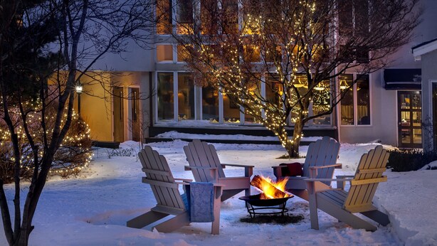 A snow-covered outdoor seating area with Adirondack chairs and a fire pit, surrounded by a house with illuminated windows and a tree with lights. - The Orchards Hotel (Williamstown)