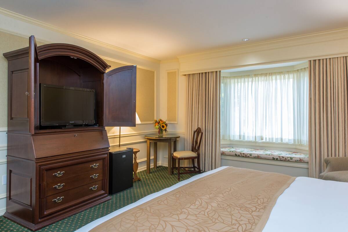 A hotel room with a large bed, a wooden dresser with drawers, a television, a small table with a lamp, a chair, and a window with curtains.