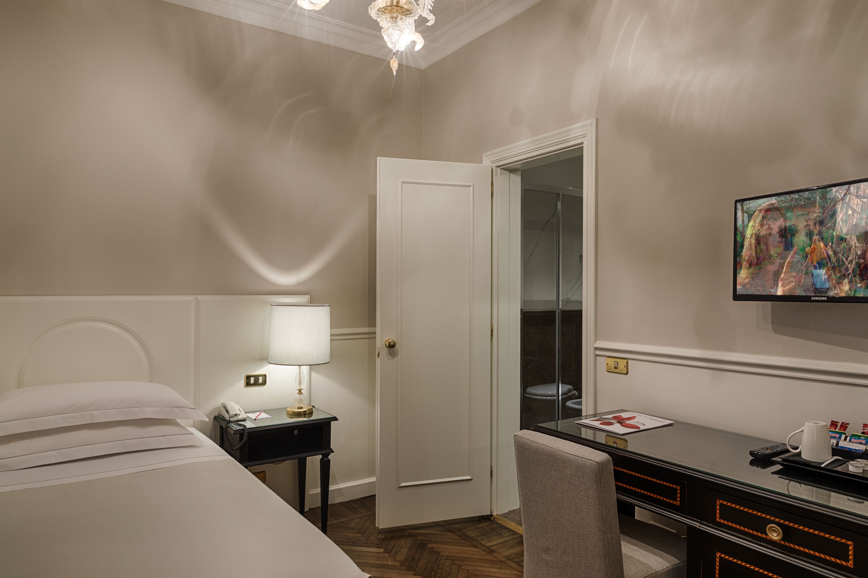 A hotel room with a bed, a desk, a chair, a lamp, a painting, and a door.