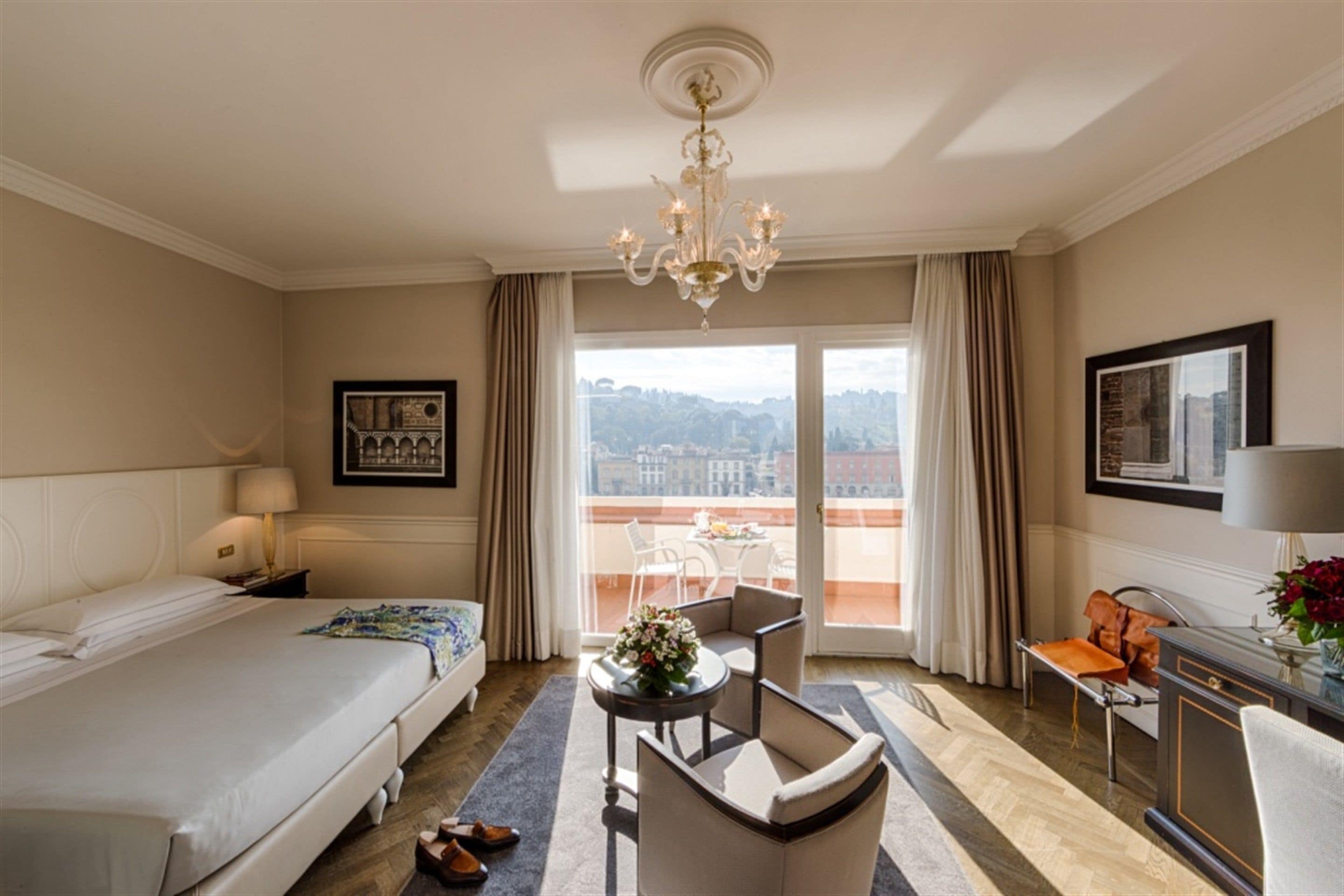 Habitación de hotel con una cama, un escritorio, una silla, una mesa con un florero y vistas a la ciudad a través de la ventana.