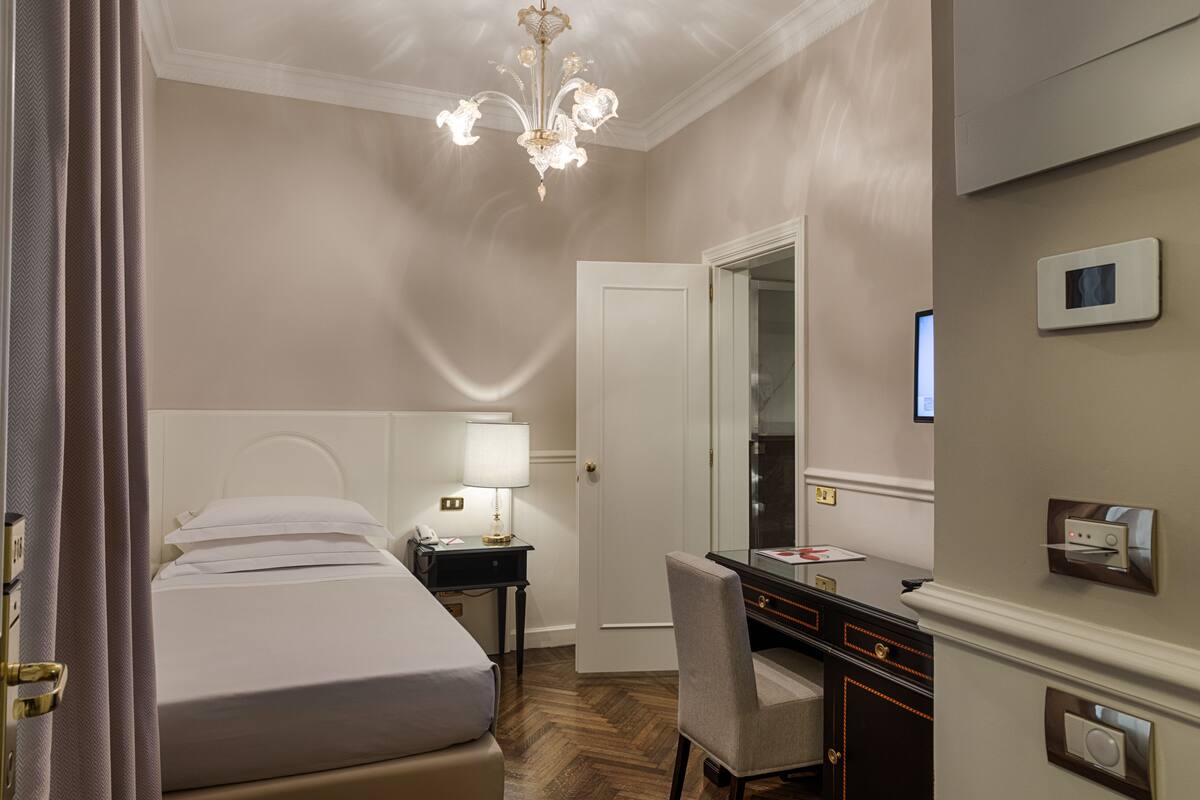 A hotel room with a bed, bedside table with a lamp, a desk with a chair, a mirror, and a thermostat on the wall.