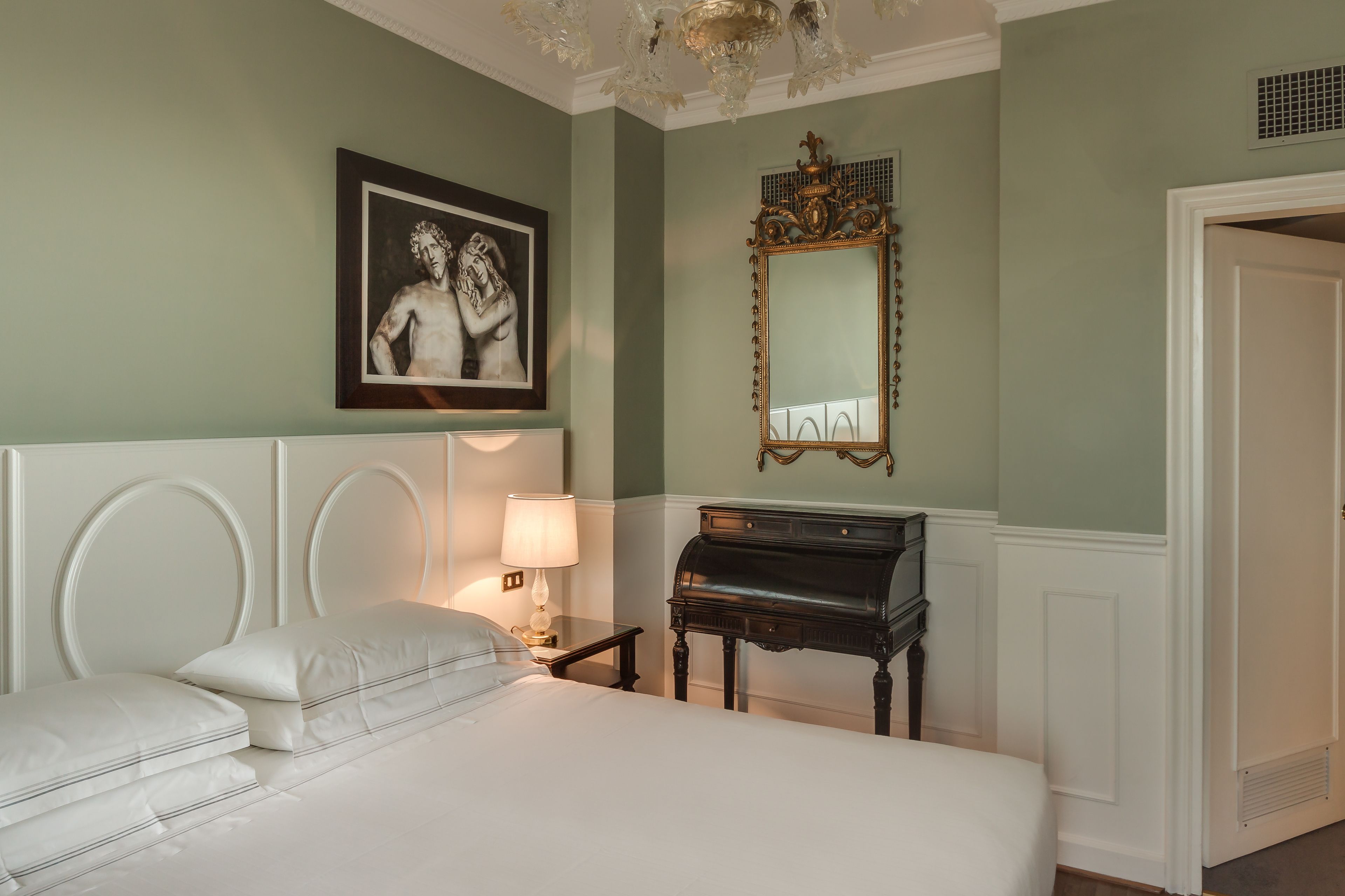 Een hotelkamer met een bed, een nachtkastje met een lamp, een piano, een lijst met foto aan de muur en een spiegel.