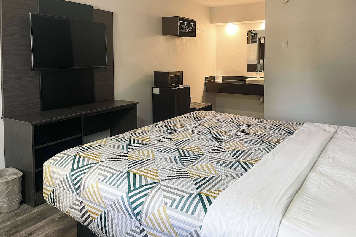 A hotel room with a bed, a TV mounted on a dark wooden cabinet, a small refrigerator, a sink with a mirror, and a trash bin.