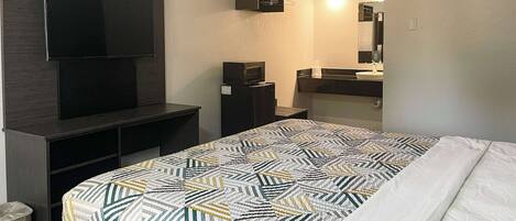 A hotel room with a bed, a TV mounted on a dark wooden cabinet, a small refrigerator, a sink with a mirror, and a trash bin.