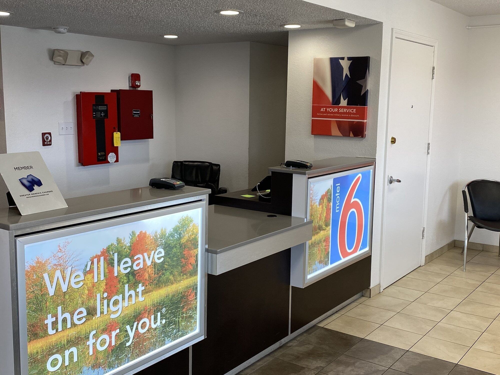 En hotellreception med en digital skärm som visar en vacker bild och text, en brandsläckare, en skylt med texten ”Medlem” och en skylt med texten ”6 Motel”.