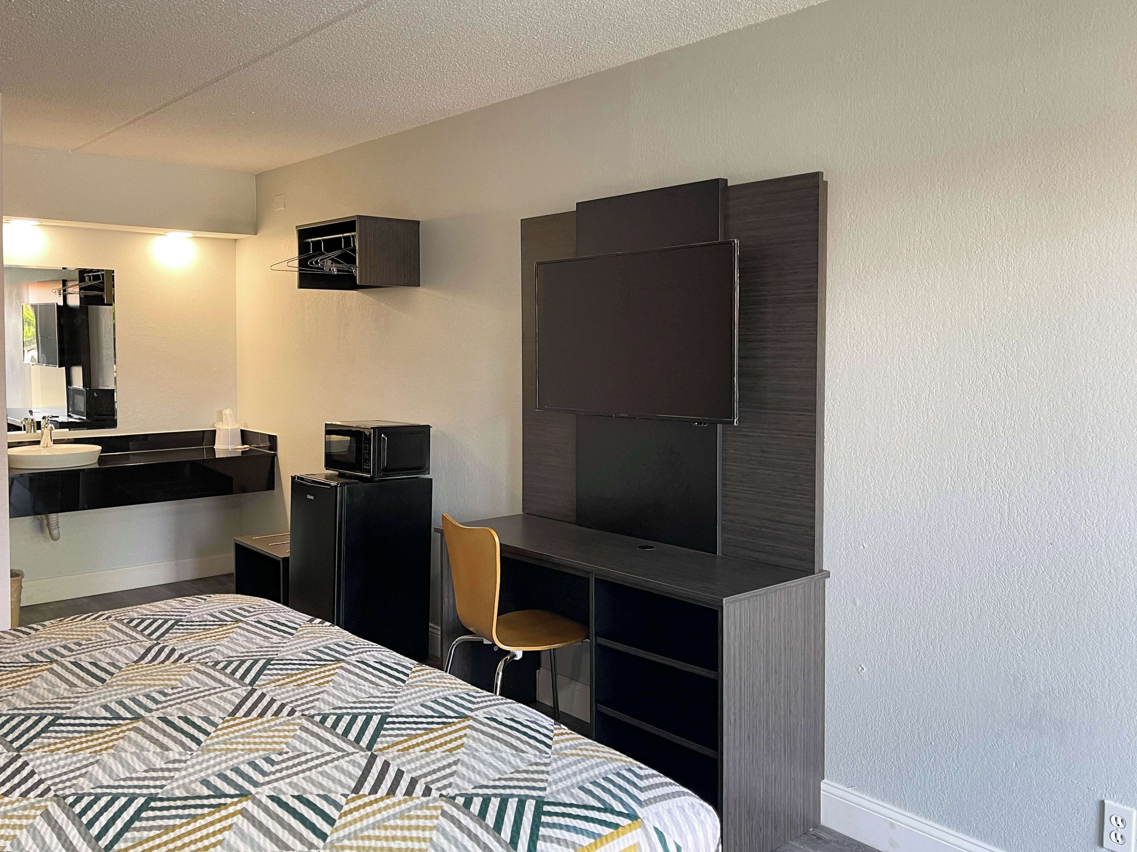 Habitación de hotel pequeña con cama, escritorio, silla, televisor, microondas y kitchenette.