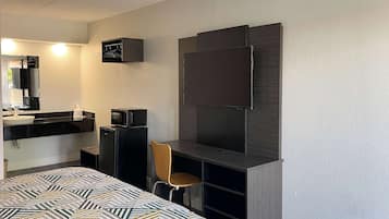 Habitación de hotel pequeña con cama, escritorio, silla, televisor, microondas y kitchenette.