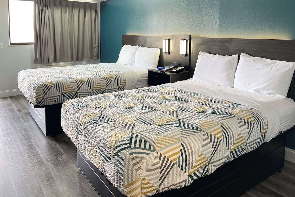 A hotel room with two beds, a nightstand, a lamp, and a window with curtains.