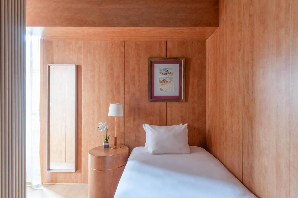 A hotel room with a wooden wall, a bed with white bedding, a round bedside table with a lamp, and a framed picture on the wall.