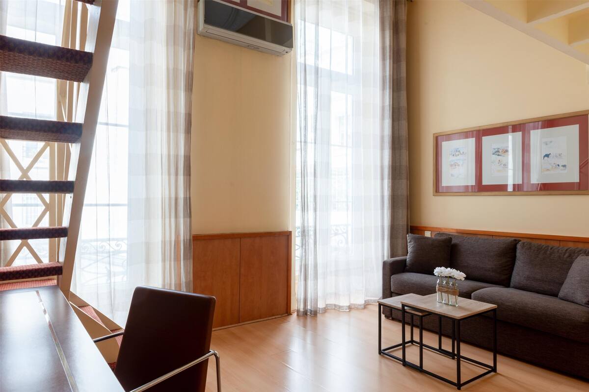 A room with a wooden floor, a sofa, a table, and a chair. There is a window with sheer curtains and a wall-mounted air conditioning unit.
