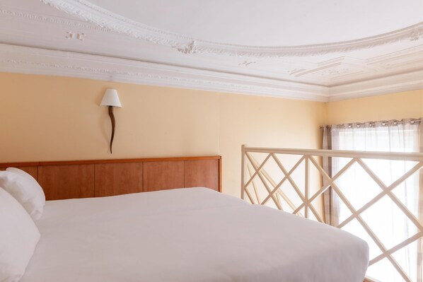 A hotel room with a wooden headboard, a white bedspread, a wall-mounted lamp, and a window with a view.