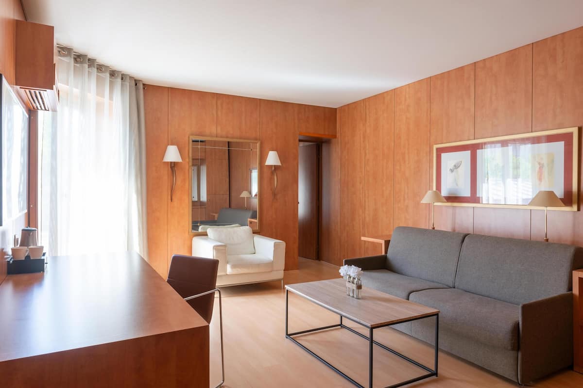 A modern living room with a wooden panel wall, a grey sofa, a white armchair, a wooden coffee table, and a large mirror.
