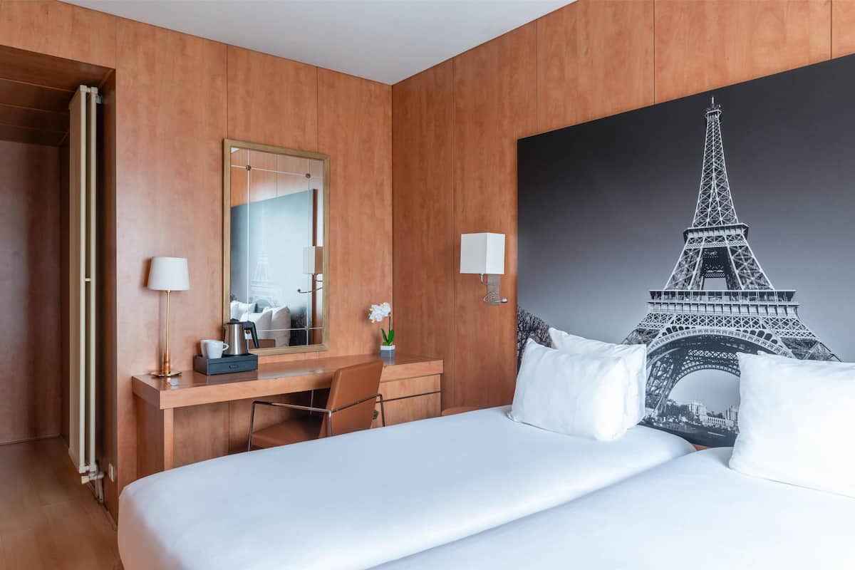 A hotel room with a bed, a desk, a chair, a mirror, a lamp, and a large picture of the Eiffel Tower.