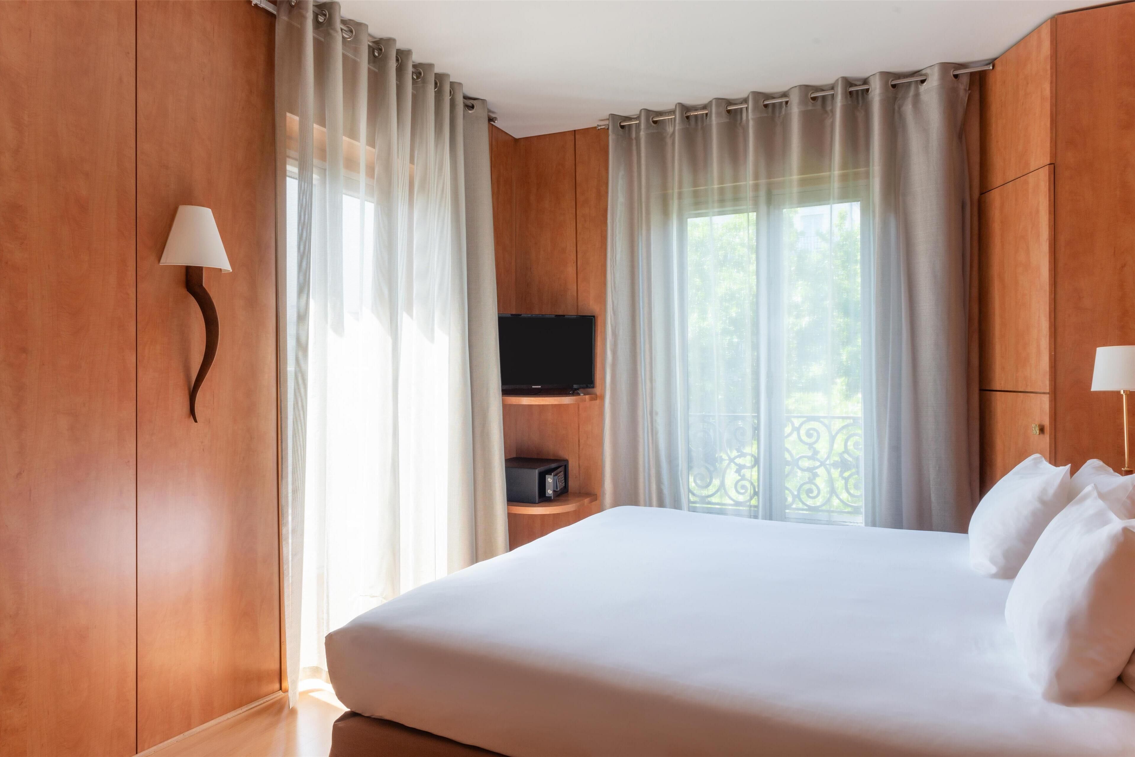 A hotel room with a large bed, a TV on a wooden shelf, and a balcony with a wrought iron railing.