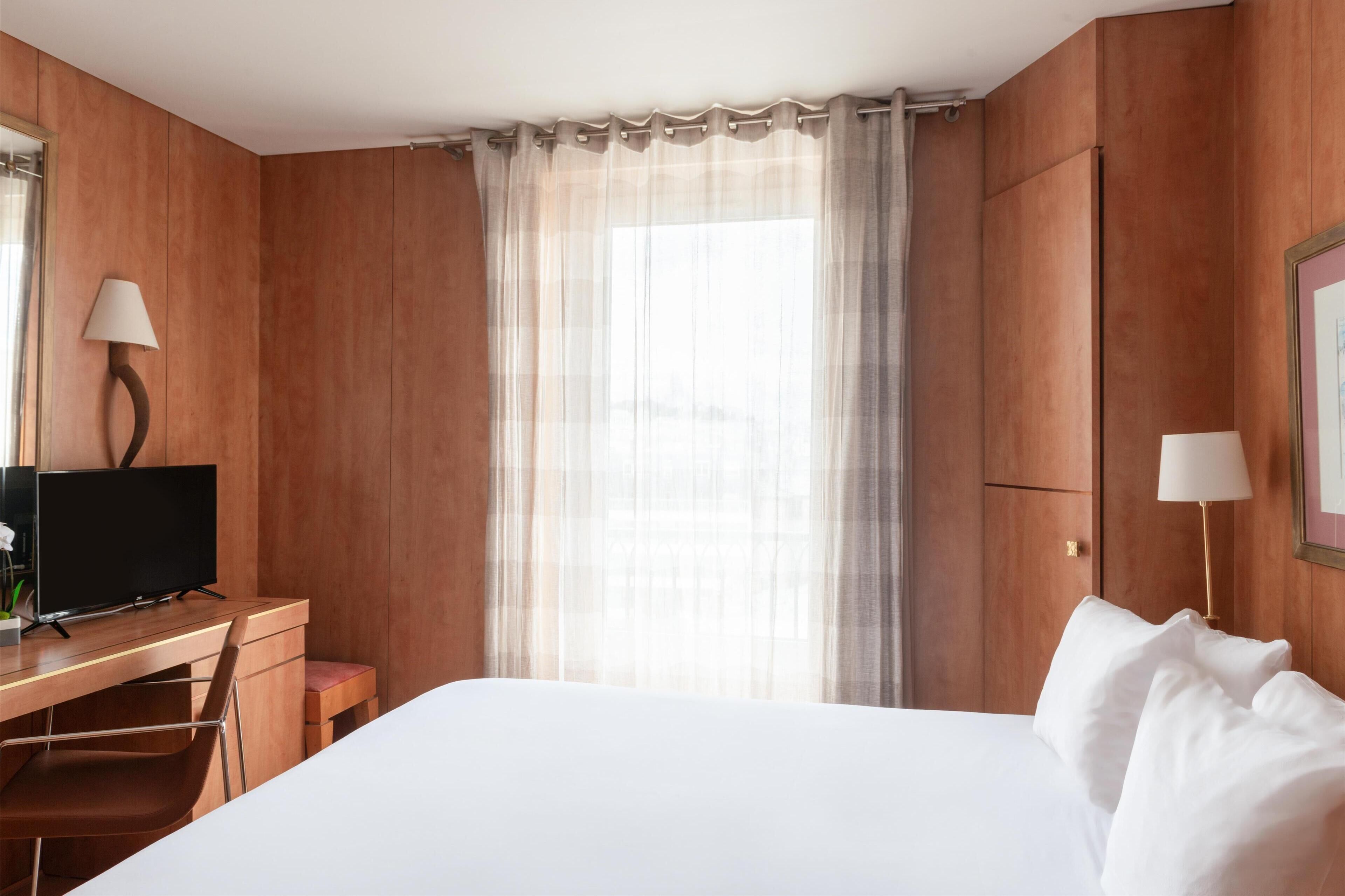 Een hotelkamer met een wand van houten panelen, een bed met witte lakens, een bureau met een televisie en een raam met dunne gordijnen.