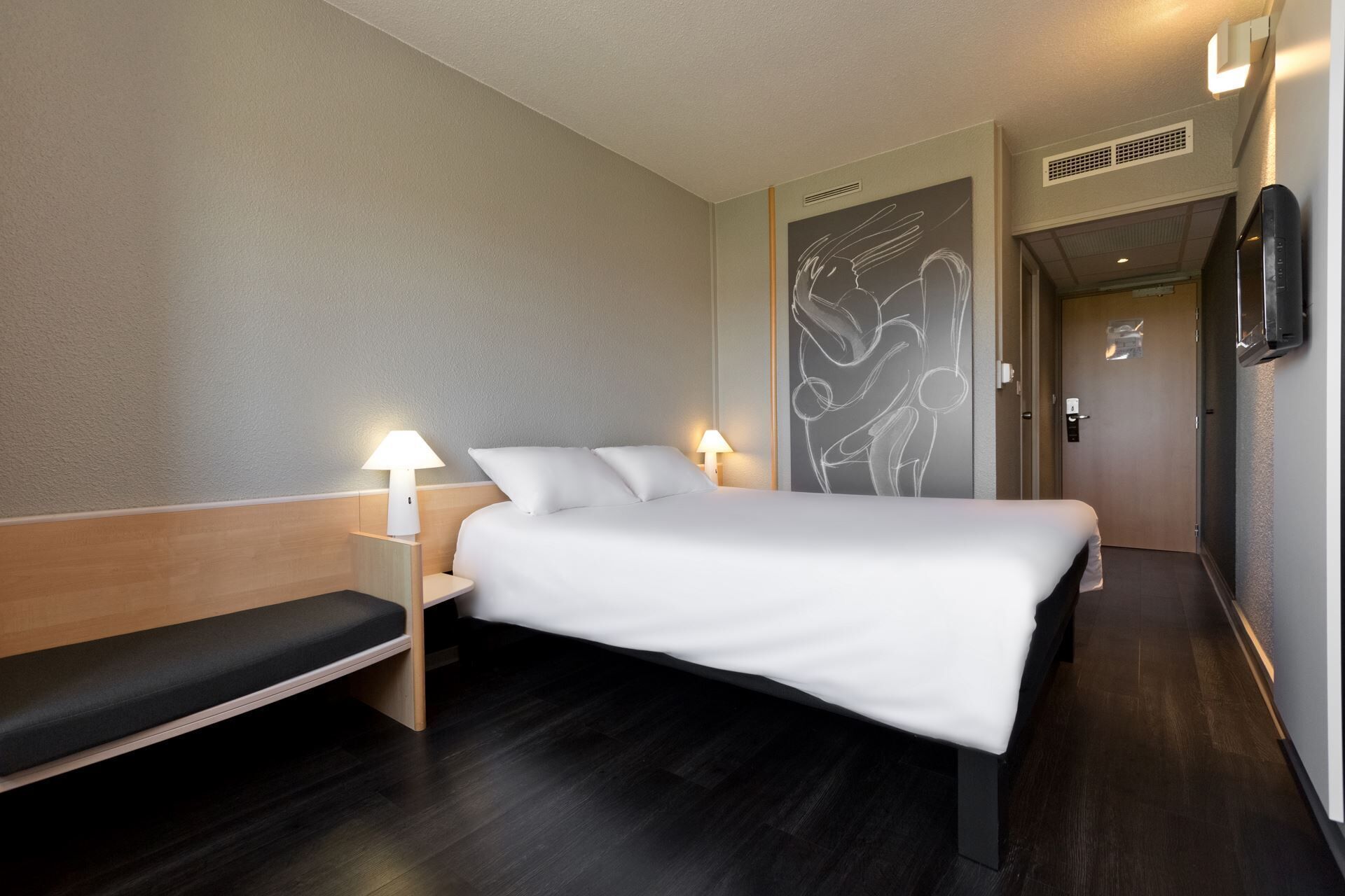 A hotel room with a bed, bedside tables, a bench, a wall with abstract art, a door, and a wall-mounted TV.