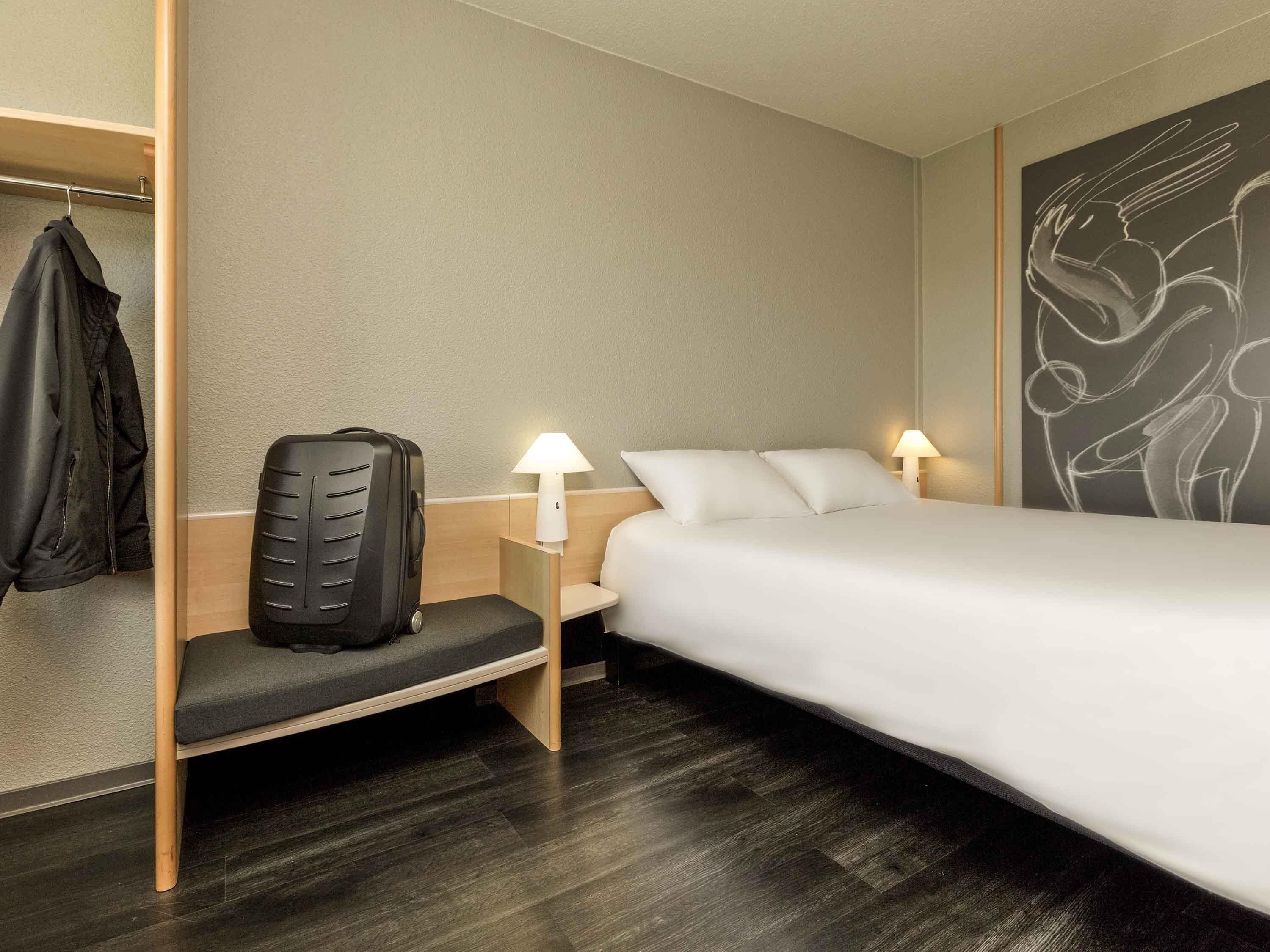 Una habitación de hotel con una cama, lámparas de noche, una silla, perchero y una obra de arte en la pared.