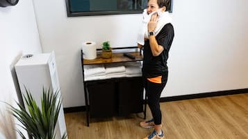A person standing in a room with a white wall, a wooden floor, a small table with a plant, a roll of toilet paper, and a white appliance.