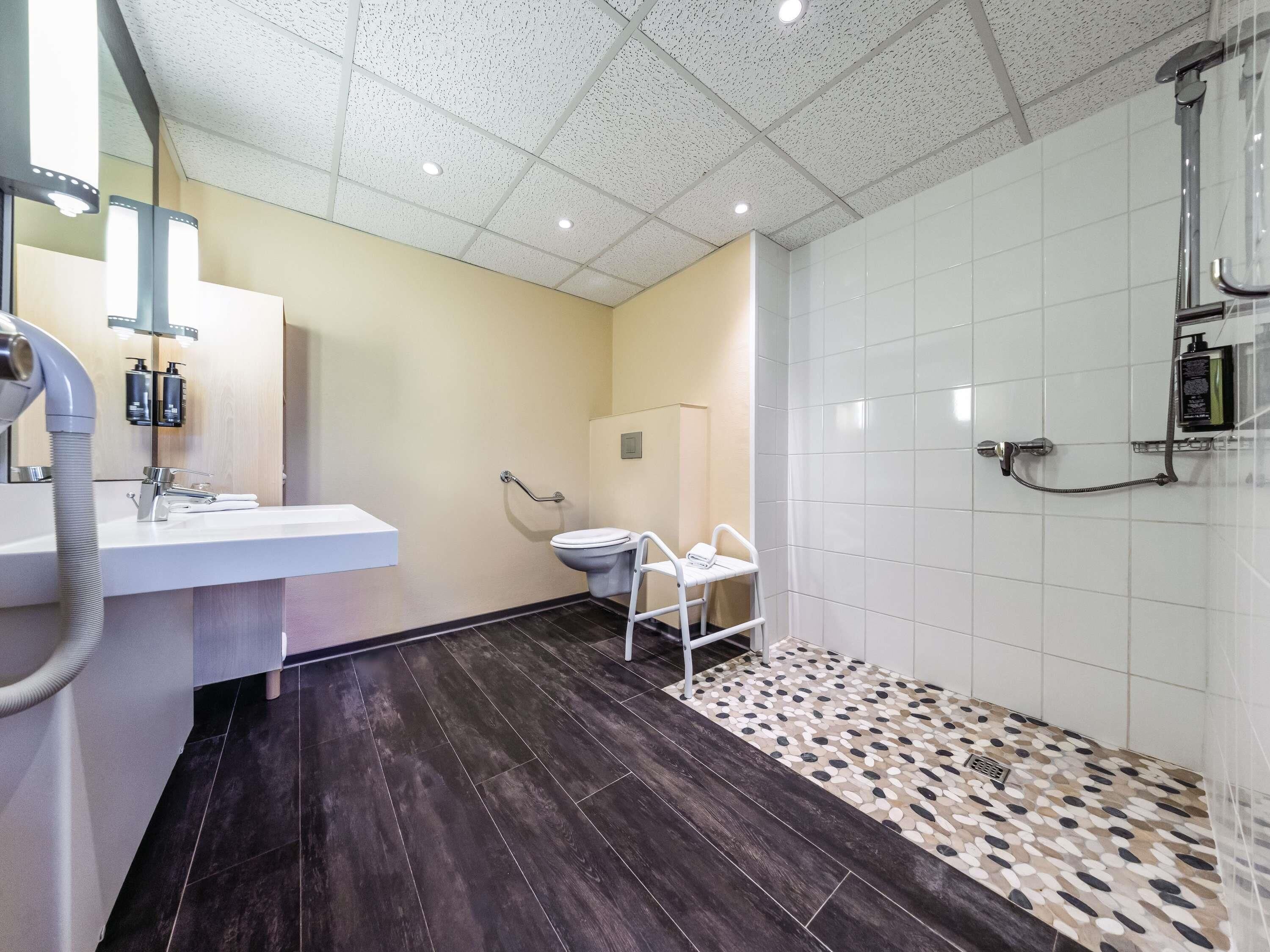 A modern bathroom with a white sink, a toilet with a grab bar, a shower area with a handheld showerhead, and a patterned floor.