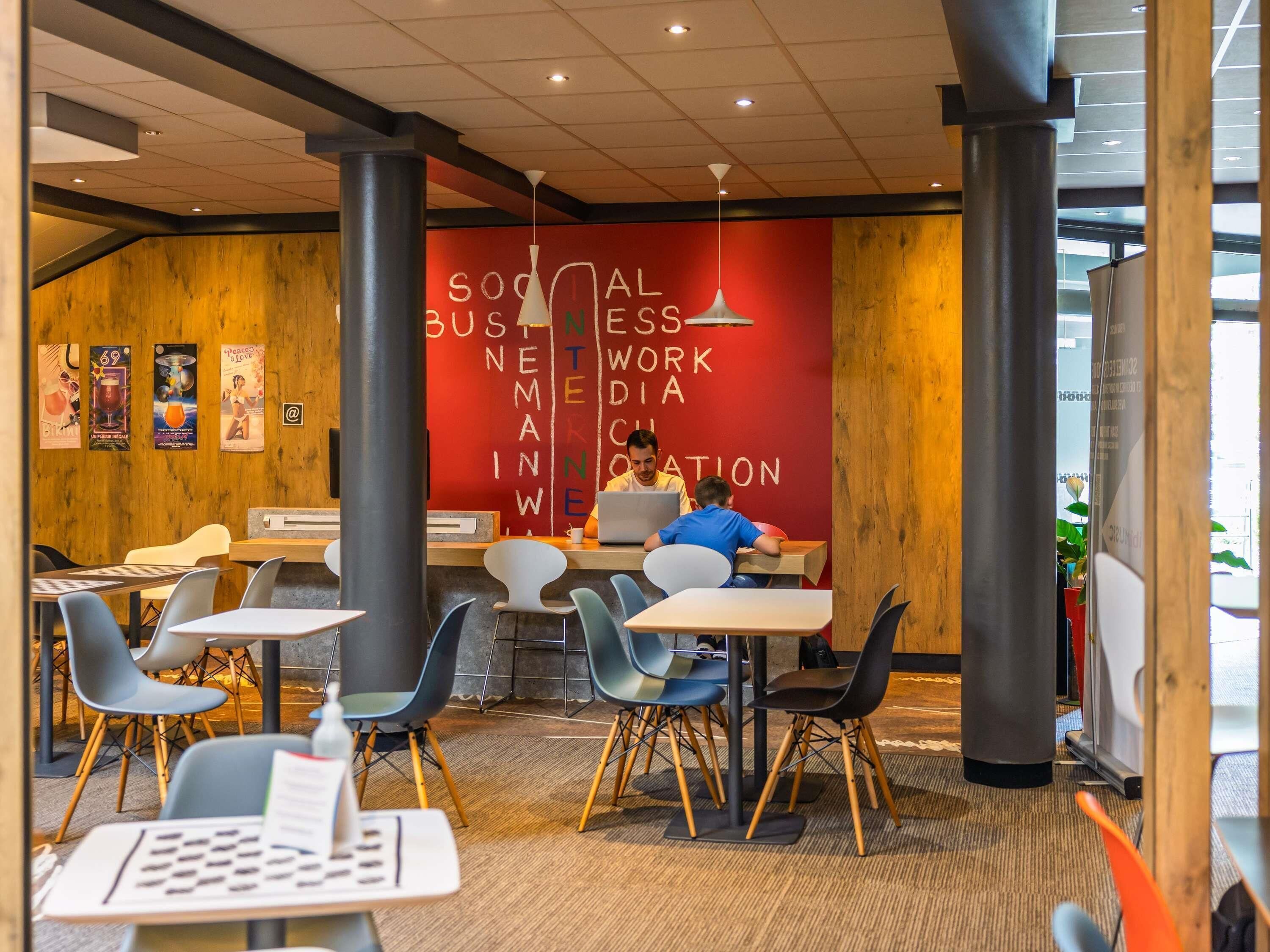 A modern workspace with a red wall featuring the text 'SOCIAL BUSINESS NETWORK MEDIA INNOVATION.' There are multiple tables and chairs, and two individuals are working at a table with laptops.