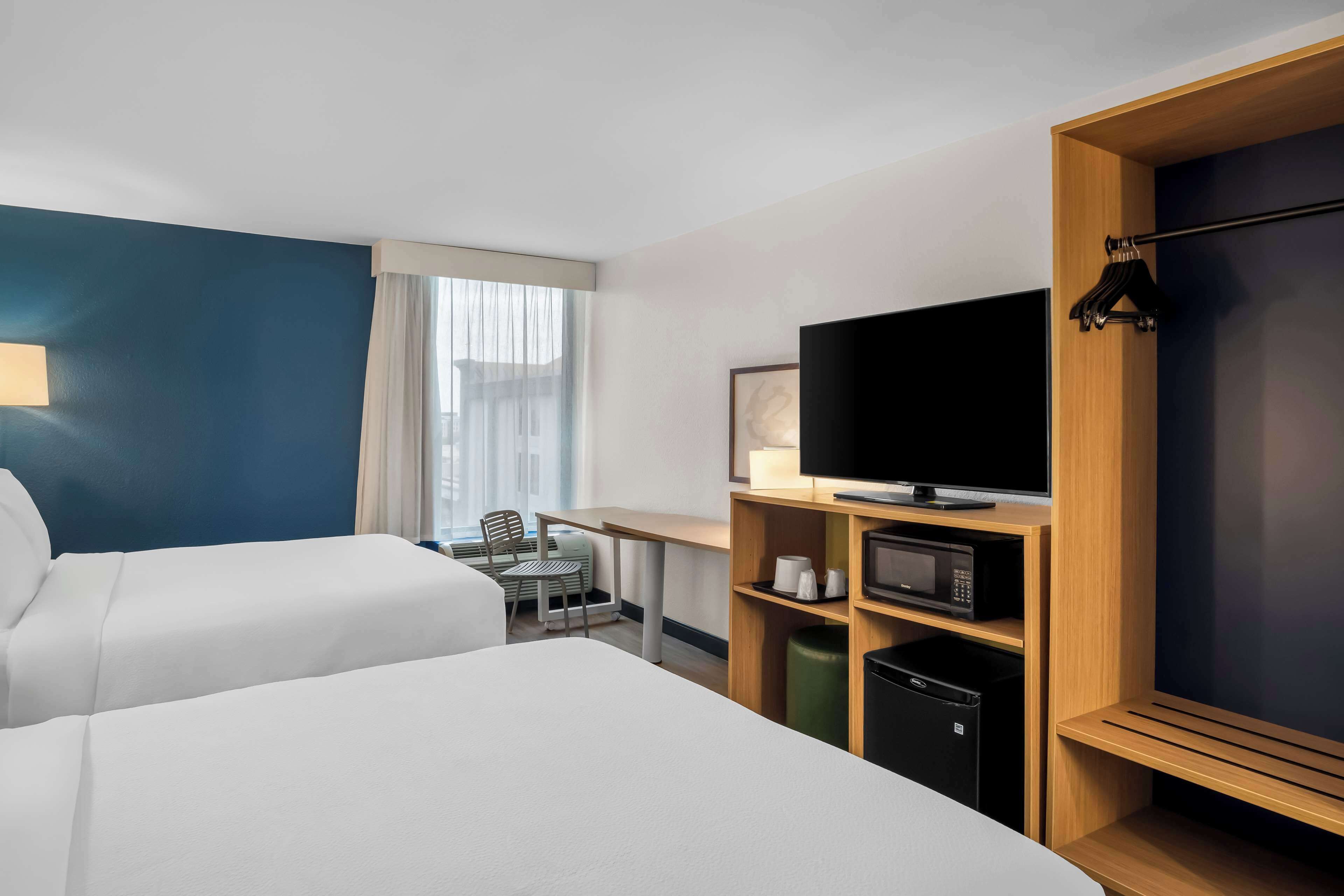 Quarto de hotel com duas camas, uma escrivaninha com cadeira, televisão e uma pequena cozinha equipada.