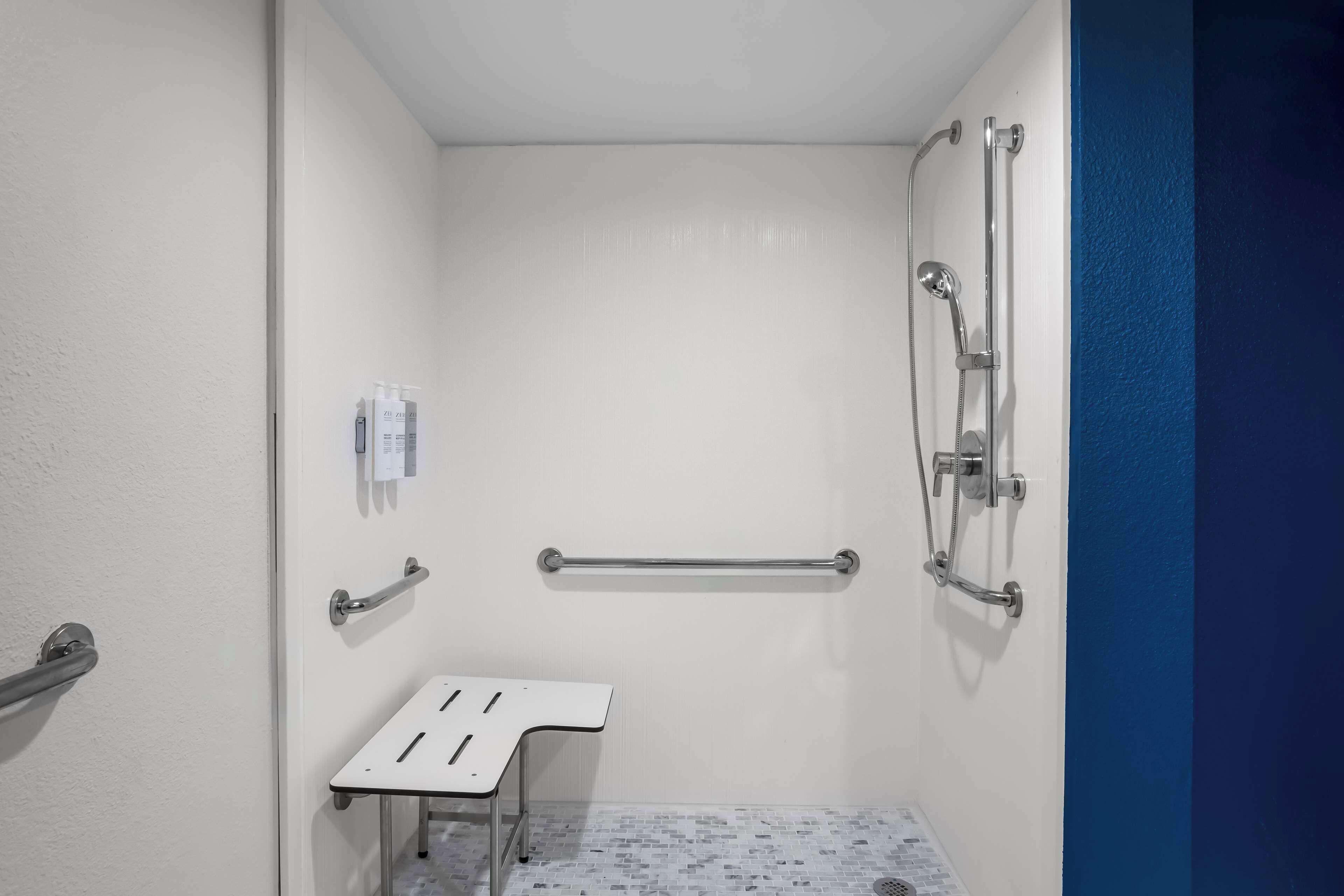 Un petit espace de douche fermé, de couleur blanche, équipé d’un banc, de barres d’appui et d’un pommeau de douche à main.