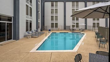 Una piscina rectangular con solárium alicatado, rodeada de sillones y mesas, y un edificio con grandes ventanales al fondo.