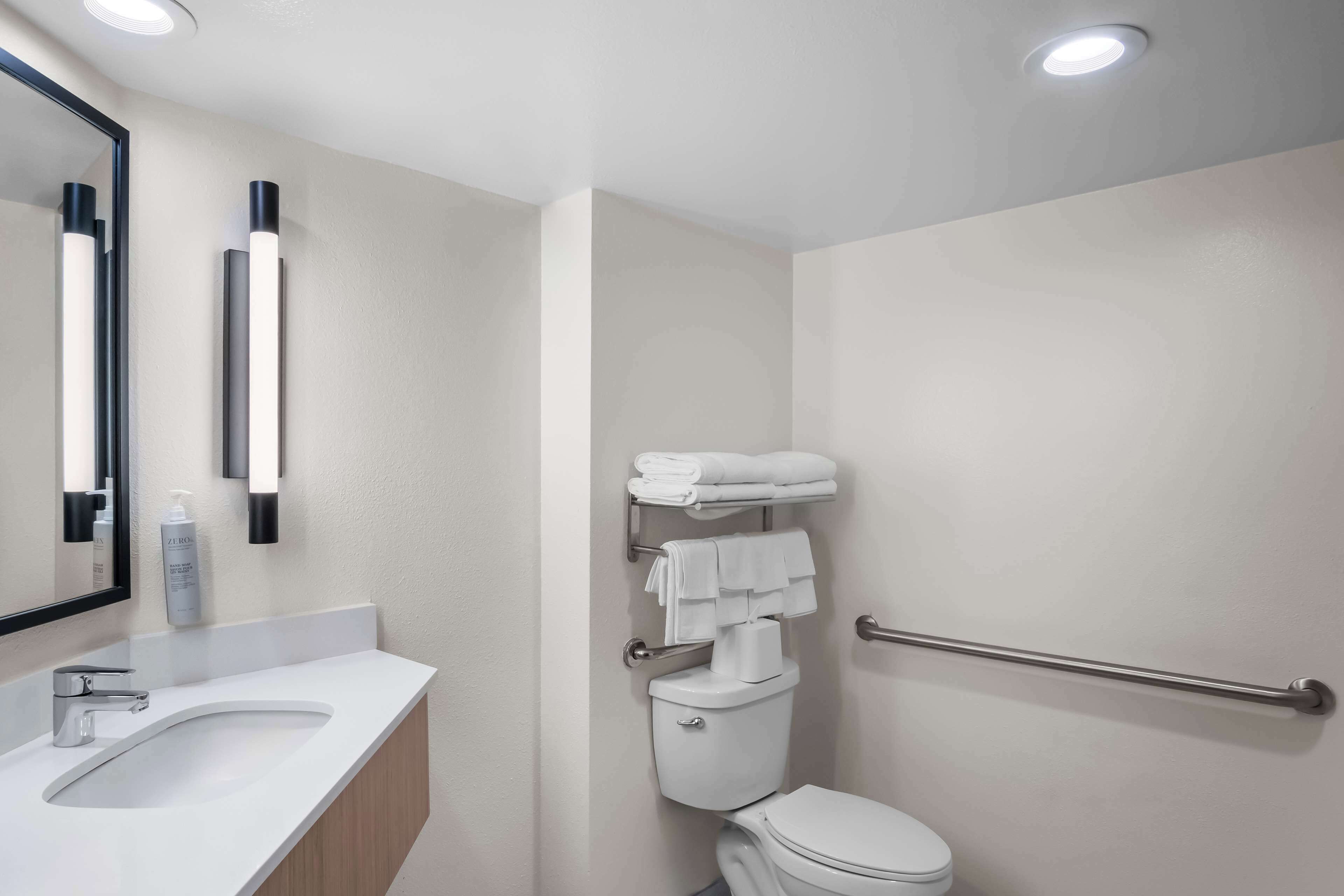 A modern bathroom with a white sink, mirror, and toilet. There are two wall-mounted lights and a towel rack with towels.