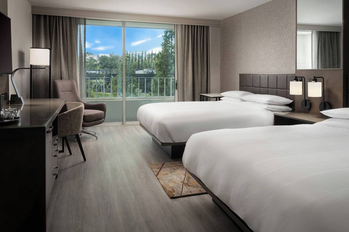 Habitación de hotel con un ventanal grande, dos camas, un escritorio, una silla y una mesita de noche con lámparas.