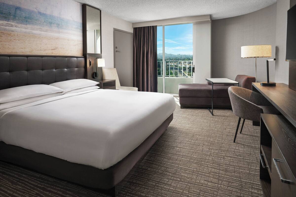 Habitación de hotel con una cama grande, un escritorio, una silla y vistas a la ciudad desde la ventana.