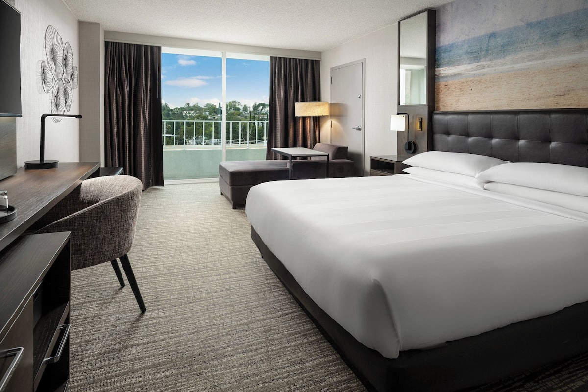 Habitación de hotel con una cama grande, un escritorio con silla, una mesita, una lámpara y vistas al exterior desde un balcón.