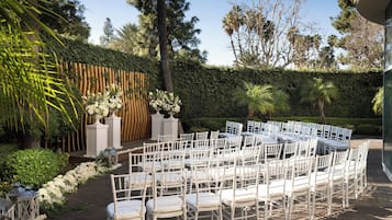 A garden setting with white chairs arranged in rows, a wooden fence, and a hedge in the background.