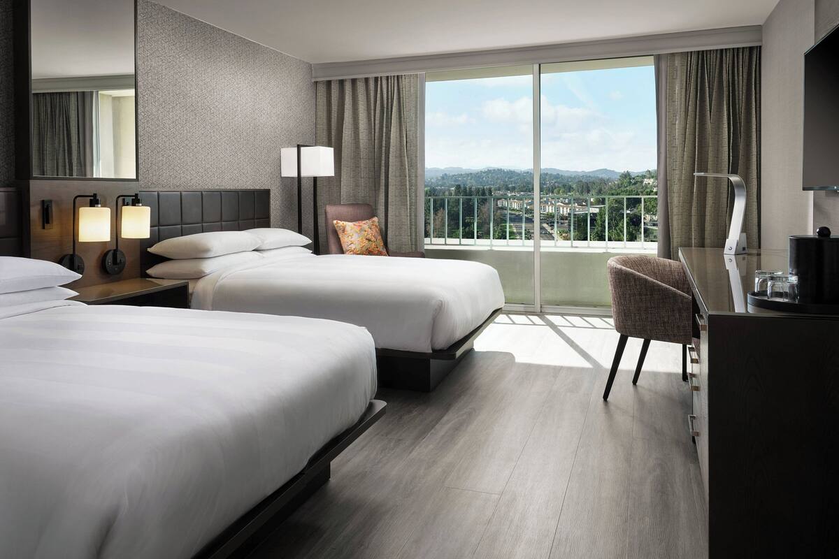 Habitación de hotel con dos camas, un escritorio, una silla y un ventanal con vista a la ciudad.