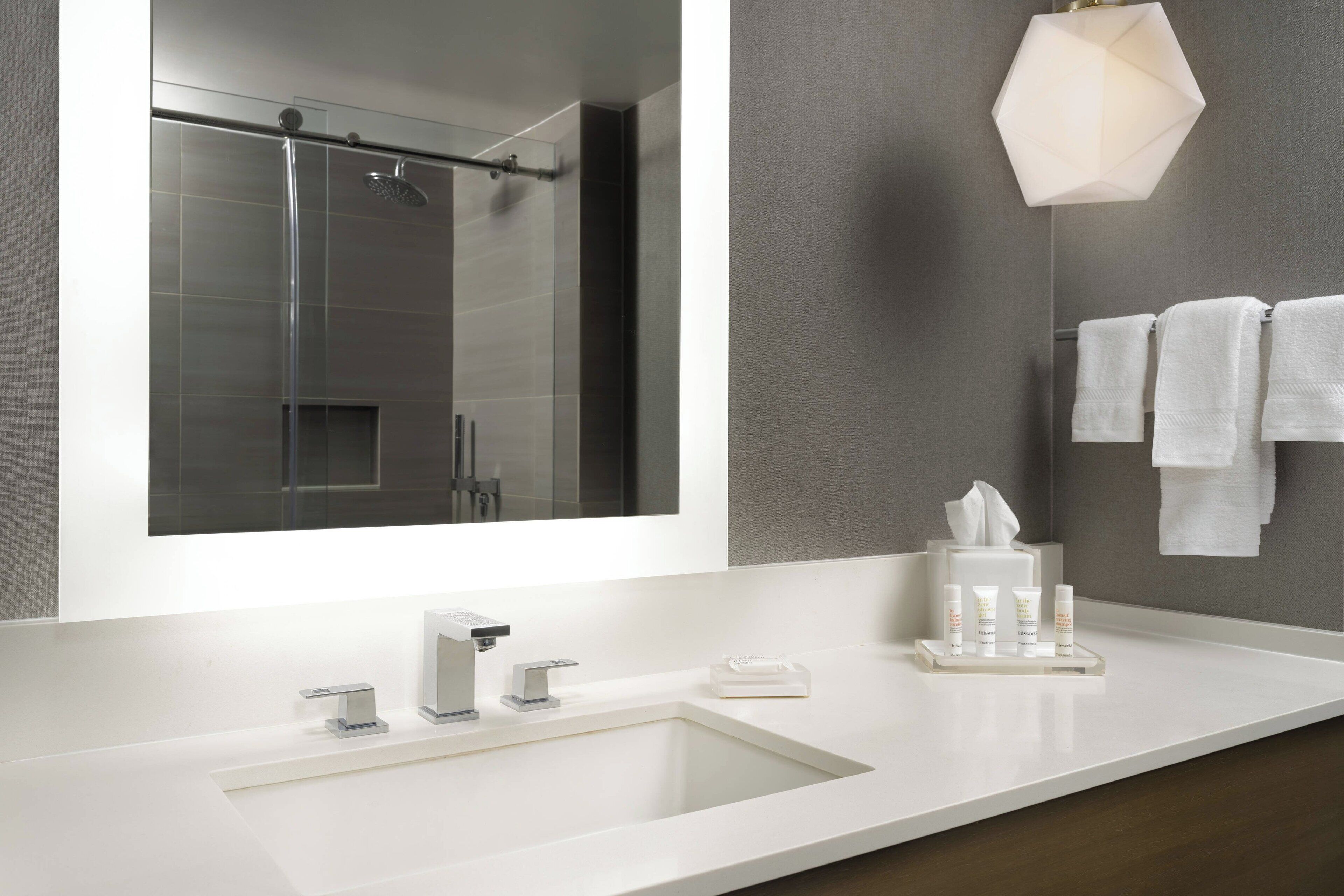 Une salle de bain moderne avec un grand miroir, une douche en verre, un lavabo blanc avec un seul robinet et un porte-serviettes avec plusieurs serviettes.