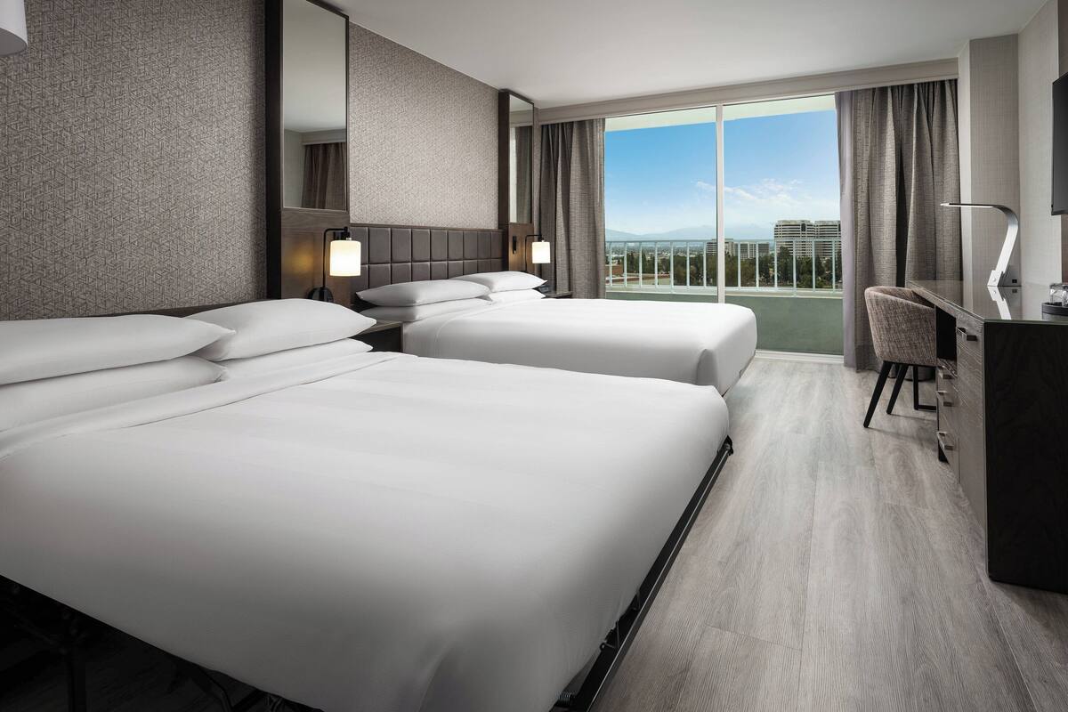 Habitación de hotel con dos camas, un escritorio, una silla, un televisor y un ventanal con vistas a la ciudad.