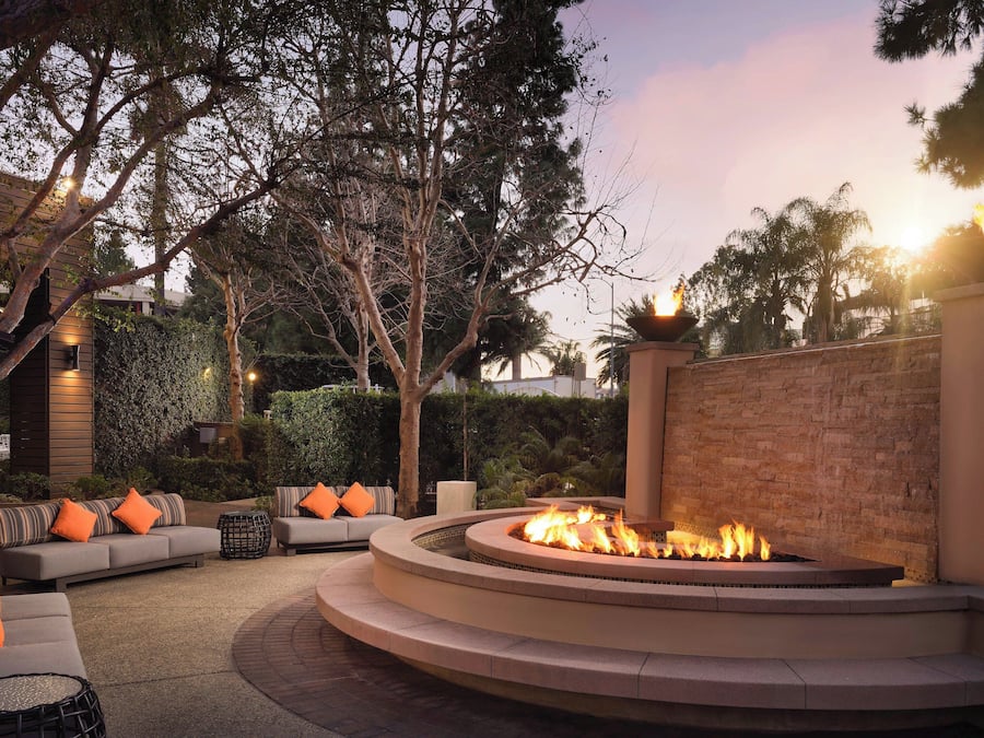 A circular outdoor seating area with a fire feature, surrounded by a low brick wall and trees.