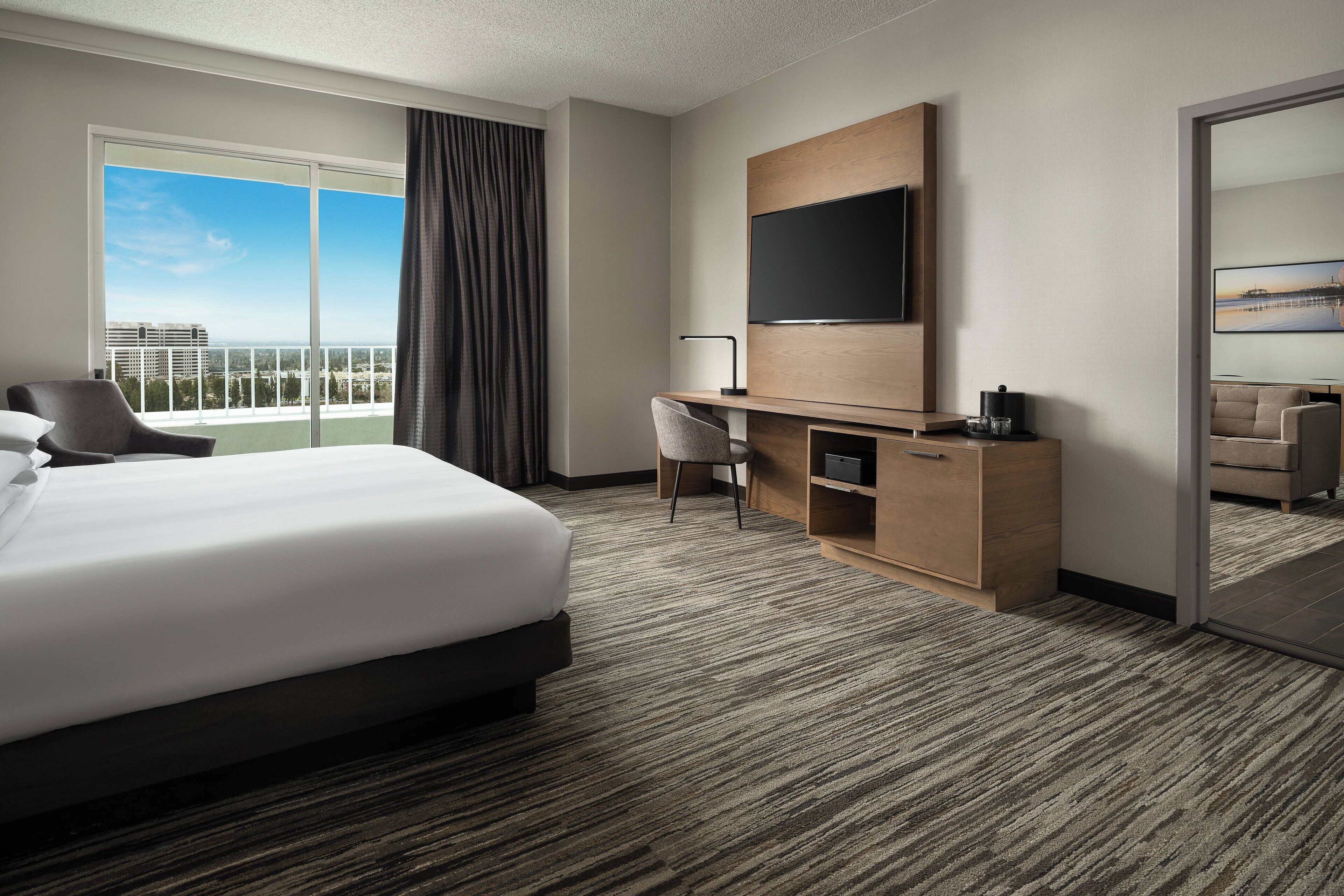 A hotel room with a large bed, a desk, a chair, a flat-screen TV, and a view of the city through a large window.