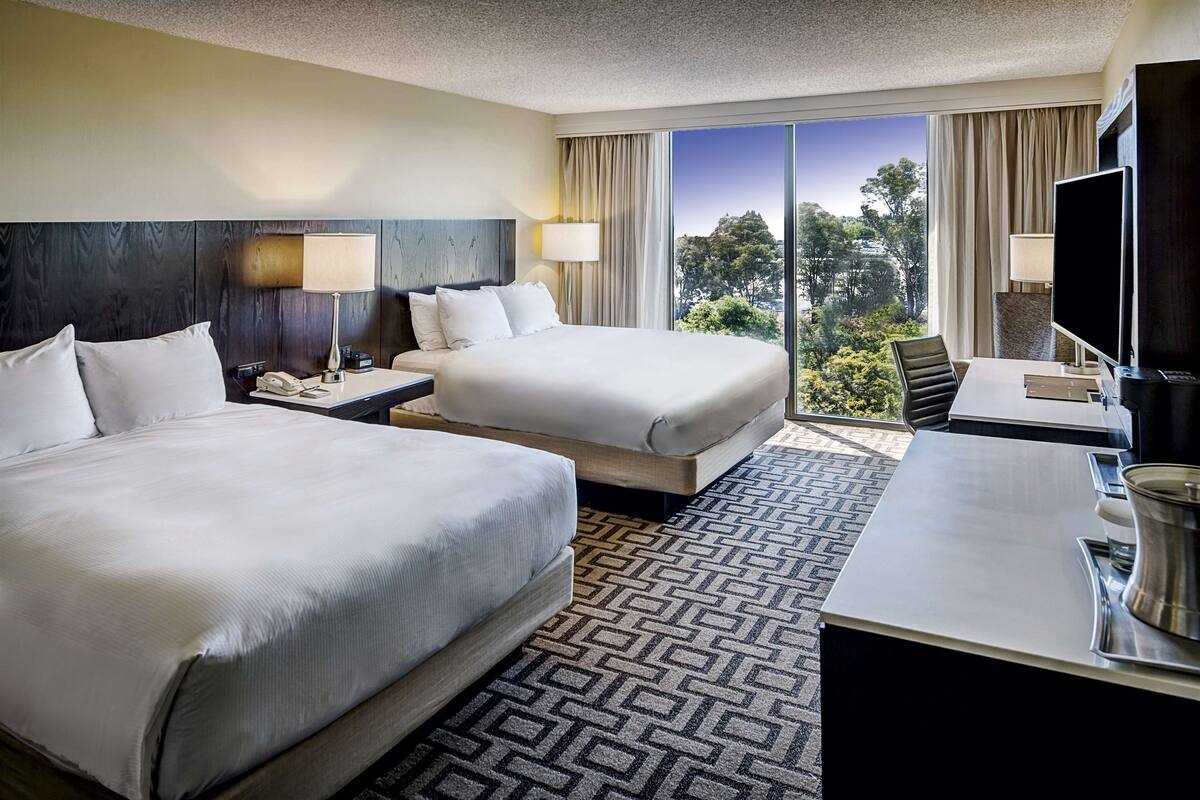 A hotel room with two beds, a desk, a TV, and a view of the outdoors.