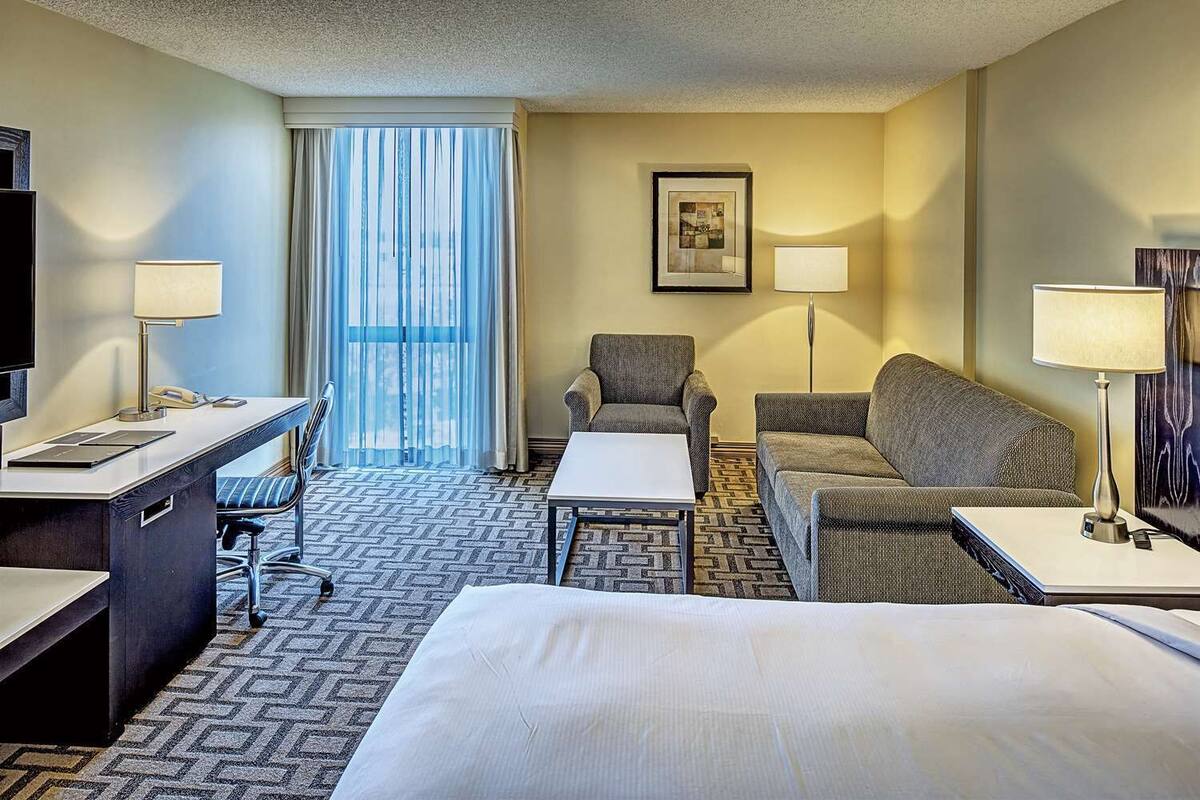 A hotel room with a bed, a desk, two armchairs, a sofa, a TV, and a window with curtains.