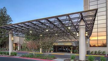 A hotel entrance with a covered walkway, a large glass facade, and a landscaped area with plants.
