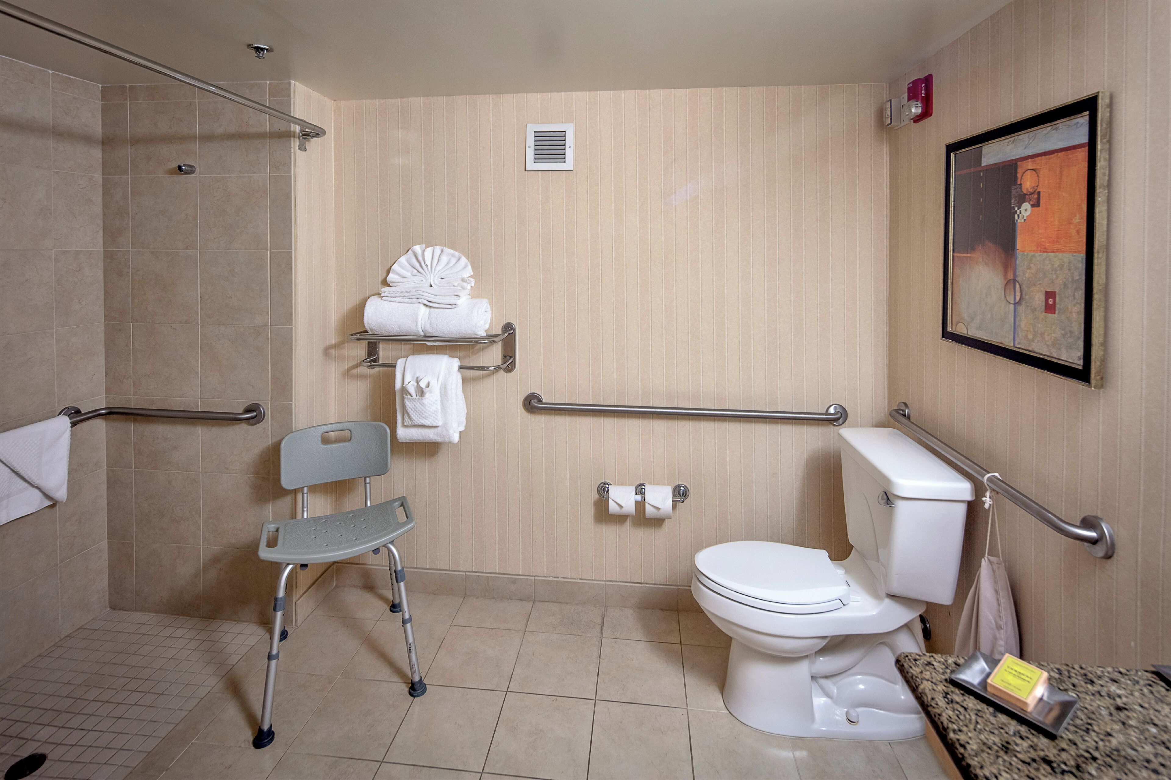 A bathroom with a shower, toilet, grab bars, a wheelchair, and a towel rack.