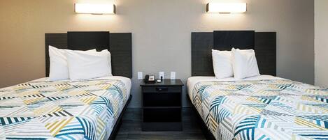 A hotel room with two beds, each with a patterned bedspread, a nightstand with a phone and a lamp, and wall-mounted lights.