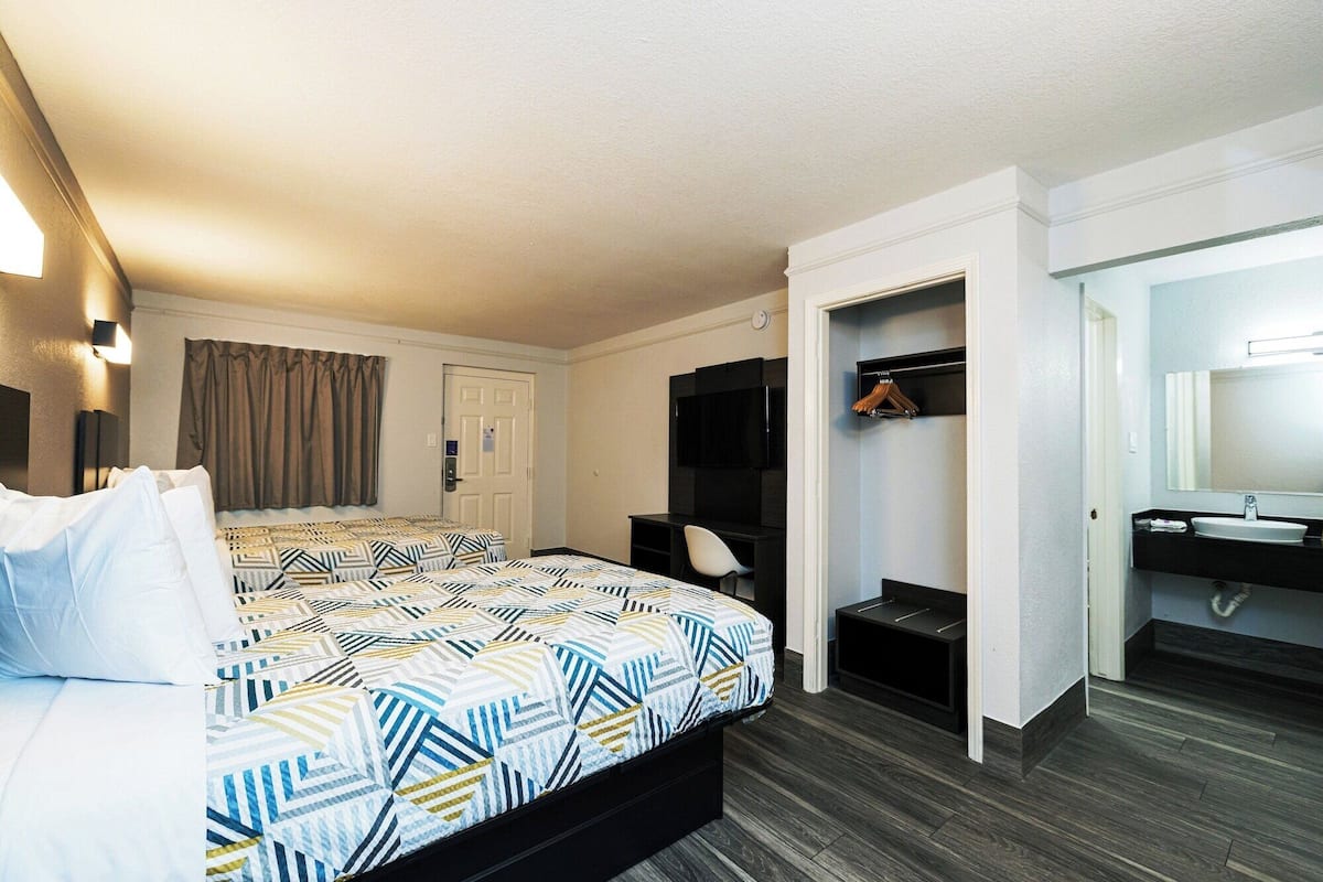 A hotel room with a bed, a desk, a chair, a TV, a wardrobe, and a bathroom with a sink and mirror.