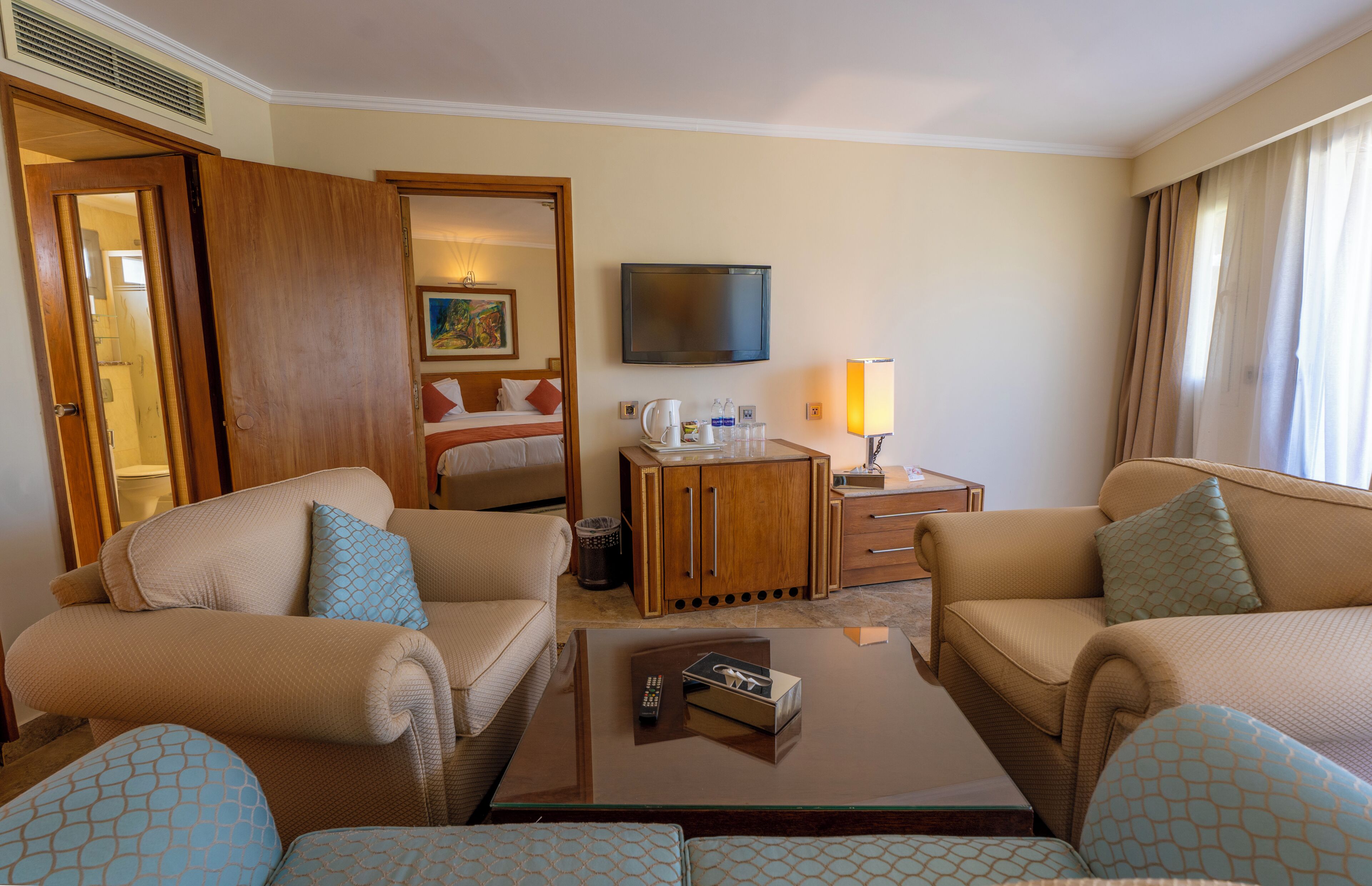 A hotel room with a sofa set, a coffee table, a TV, a bedside table with a lamp, and a door leading to a bathroom.