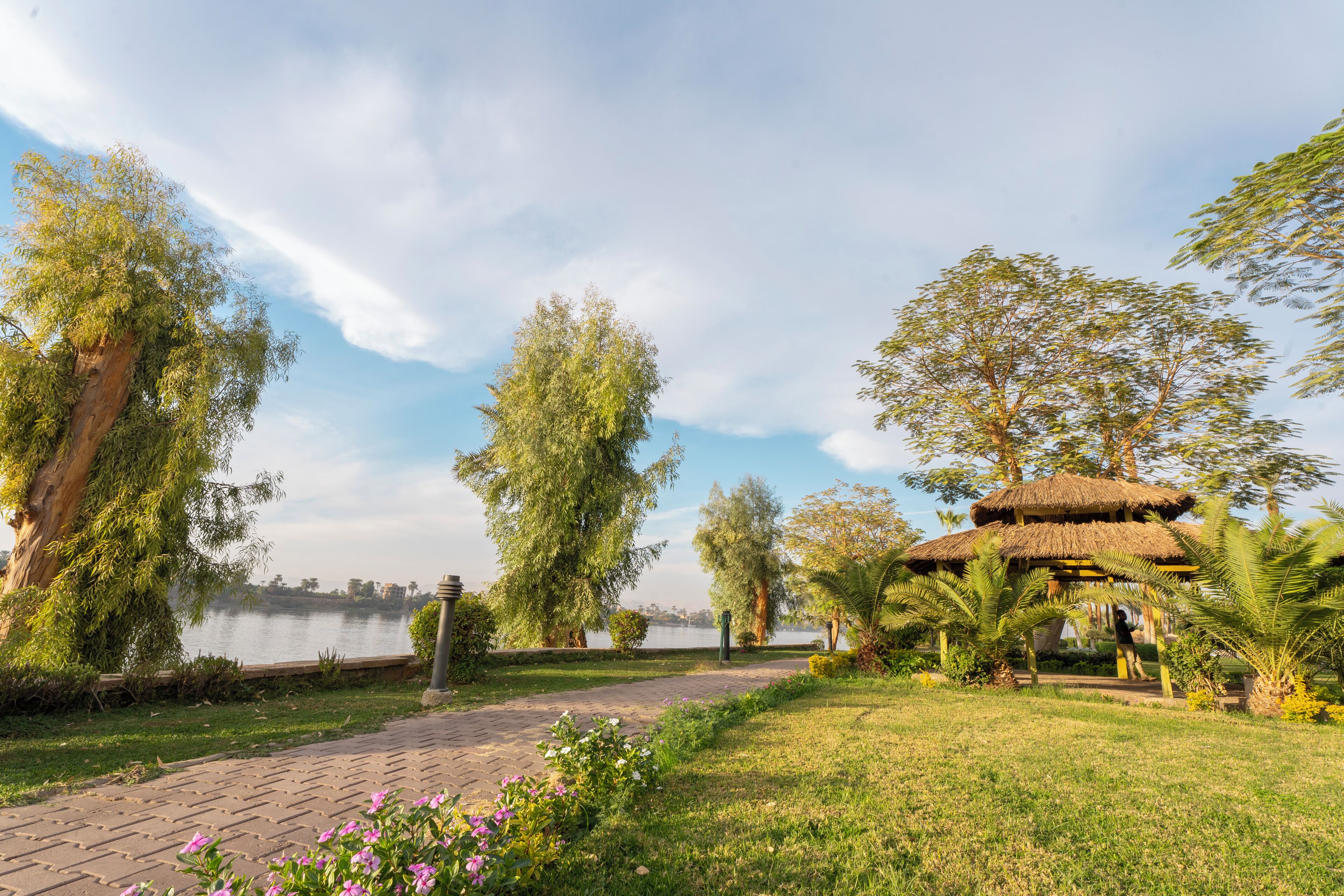 Un parc doté d’un pavillon à toit de chaume, d’une allée pavée et d’un lac en arrière-plan.