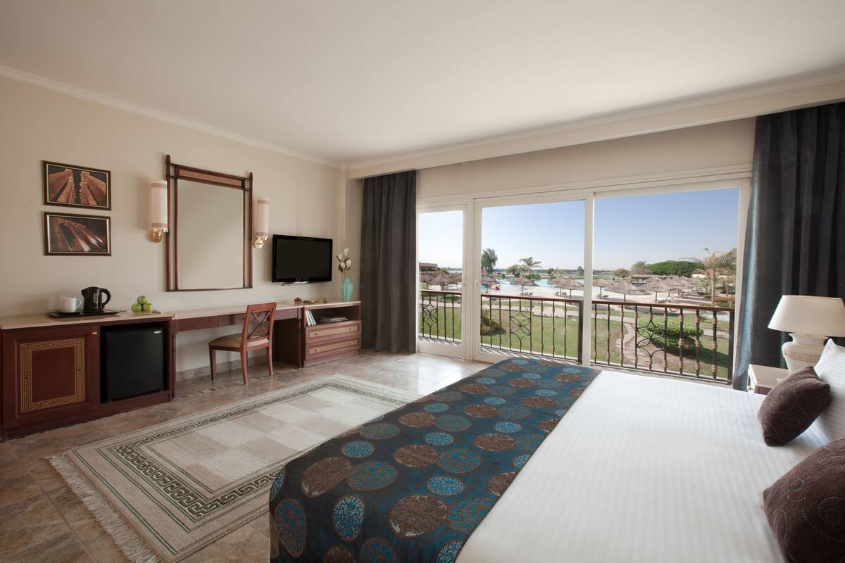 A hotel room with a large bed, a desk with a TV, a small fridge, and a balcony with a pool view.