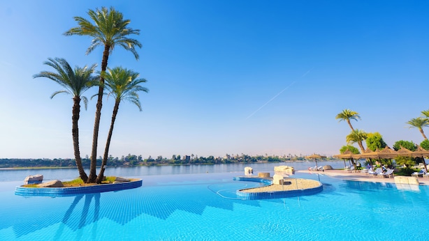 Una piscina con palmeras y sillones de descanso, rodeada de un cielo azul brillante.