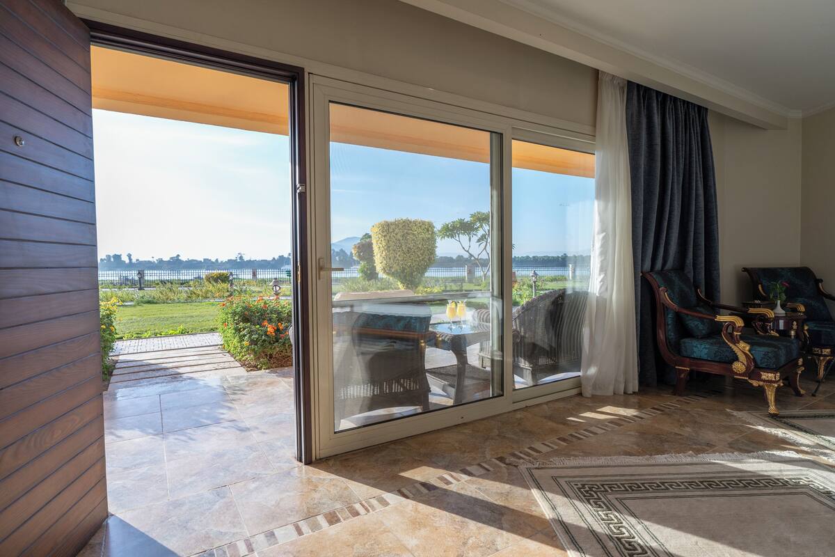 A spacious room with a sliding glass door leading to a patio, a dining area with outdoor furniture, and a view of a landscaped garden and distant hills.