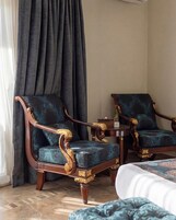 A hotel room with two ornate armchairs, a small table, and a bed with a patterned bedspread.